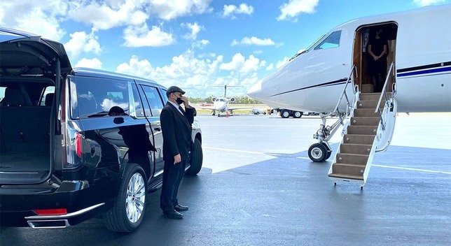 "A suited individual stands beside a black SUV with its trunk open, facing a private jet with extended stairs on an airport tarmac beneath a partly cloudy blue sky."
