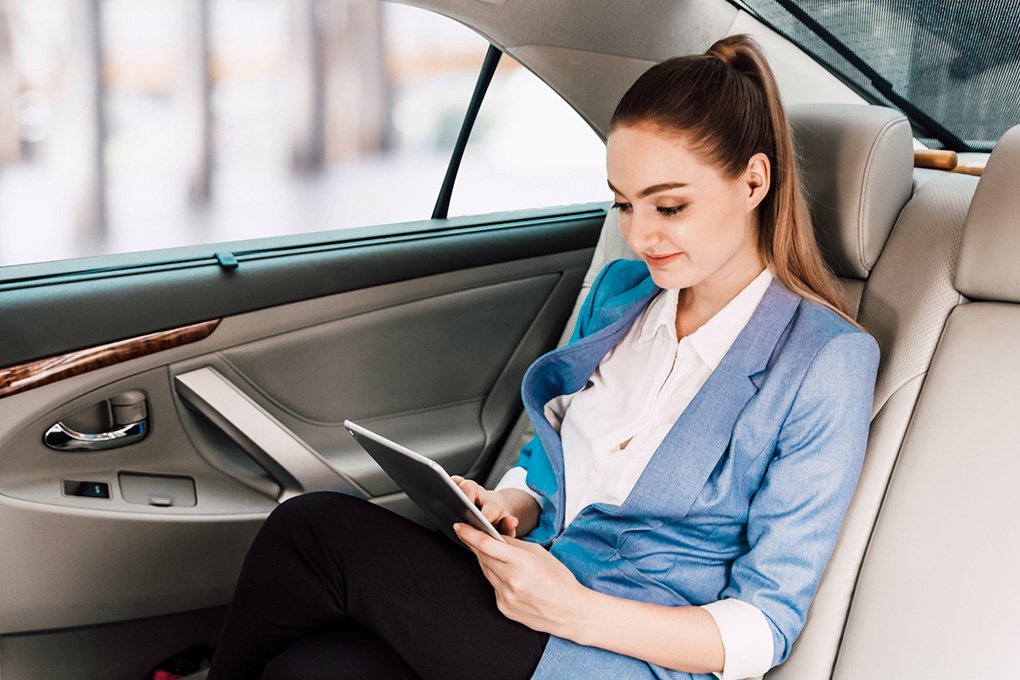 "A young woman with a ponytail, dressed in a white shirt and blue blazer, sits in the back seat of a car, focused on a tablet. The car has light-colored seats and wood-grain trim."