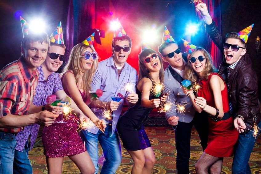 "Young adults in party hats and sunglasses celebrate with sparklers and drinks under colorful lights."