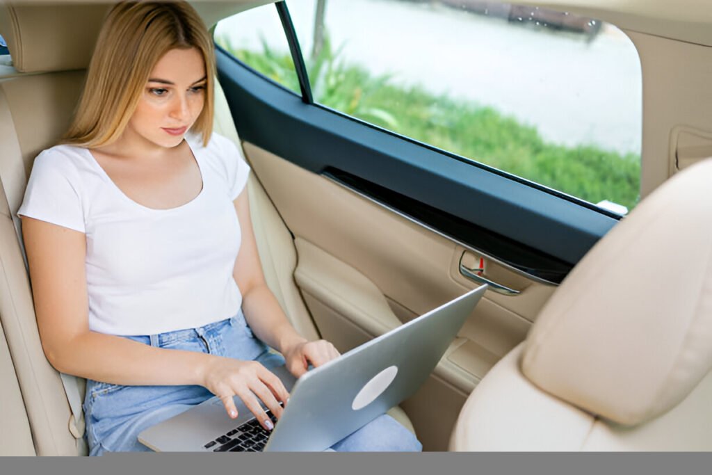"A young blonde woman in a white t-shirt and light jeans works on a silver laptop in the back seat of a light-colored car, with greenery and water visible through the window."