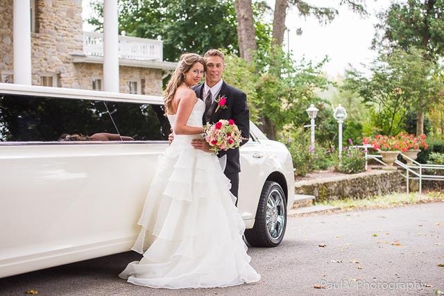 "A smiling bride in a tiered white gown embraces the groom in front of a white stretch limo, with a stone building and trees behind them."