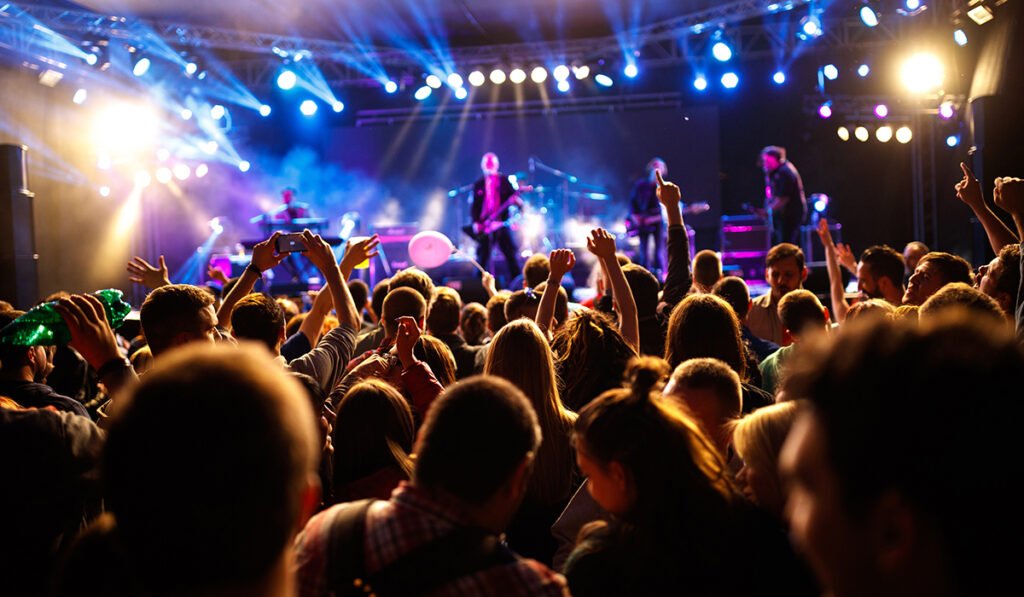 "A lively crowd at an outdoor concert or music festival, with raised hands and cell phones capturing the moment, illuminated by bright stage lights and spotlights, as a band performs on stage in the background."