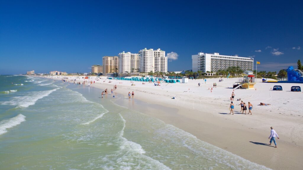 Wide shot of Clearwater Beach, Florida, with white sand, clear turquoise water, and people walking, sunbathing, or enjoying beach activities; large hotels and condos line the beachfront under a bright blue sky.