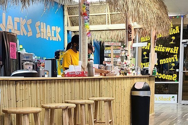 Person in yellow shirt stands behind bamboo-style counter at Jack's Shack, a dessert shop with a thatched roof, blue wall with "JACK'S SHACK" signage, wooden stools, and flavor signs like "STRAWBERRY," "PINEAPPLE," and "COCONUT."
