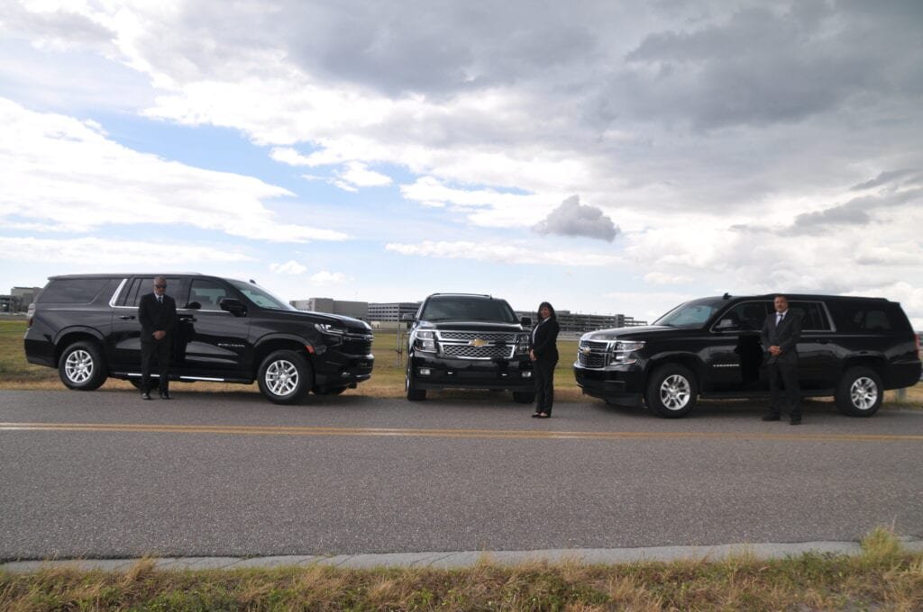 "Three dark SUVs, likely Chevrolet Suburbans, are parked on a paved lot, with two individuals in black suits standing beside them. A large building looms in the distance under a cloudy sky."