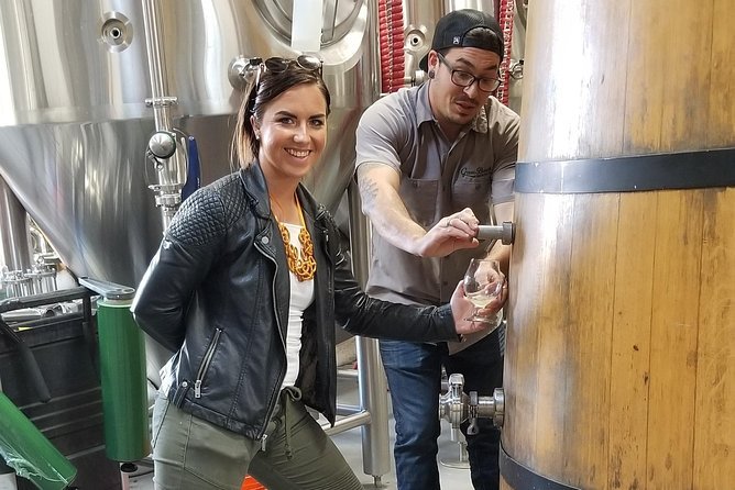 "A woman in a black leather jacket and olive pants smiles at the camera while a man in a grey shirt and black cap pours a drink from a large wooden barrel into her glass inside a brewery, with stainless steel brewing tanks in the background."