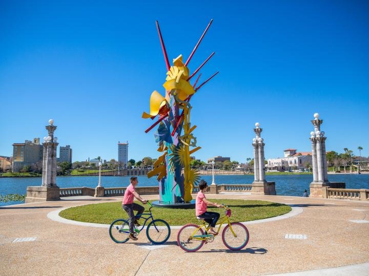 Colorful abstract metal sculpture "Tribute to the Volunteer Spirit" by Albert Paley stands on Lake Mirror's eastern shore in Lakeland, Florida; two cyclists ride past on a paved path with the lake and city skyline in the background under a clear blue sky.