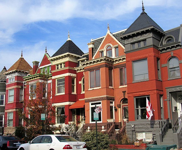 Row of vibrant, multi-colored Victorian-style houses with varied architecture and rooflines under a bright blue sky; white car parked in front.