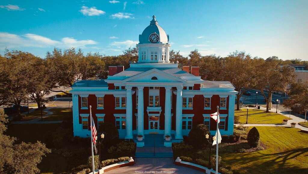 Aerial view of the historic Pasco County Courthouse in Dade City, Florida, with red brick exterior, white columns, domed clock tower, green lawns, trees, and two flags in the foreground under a blue sky.
