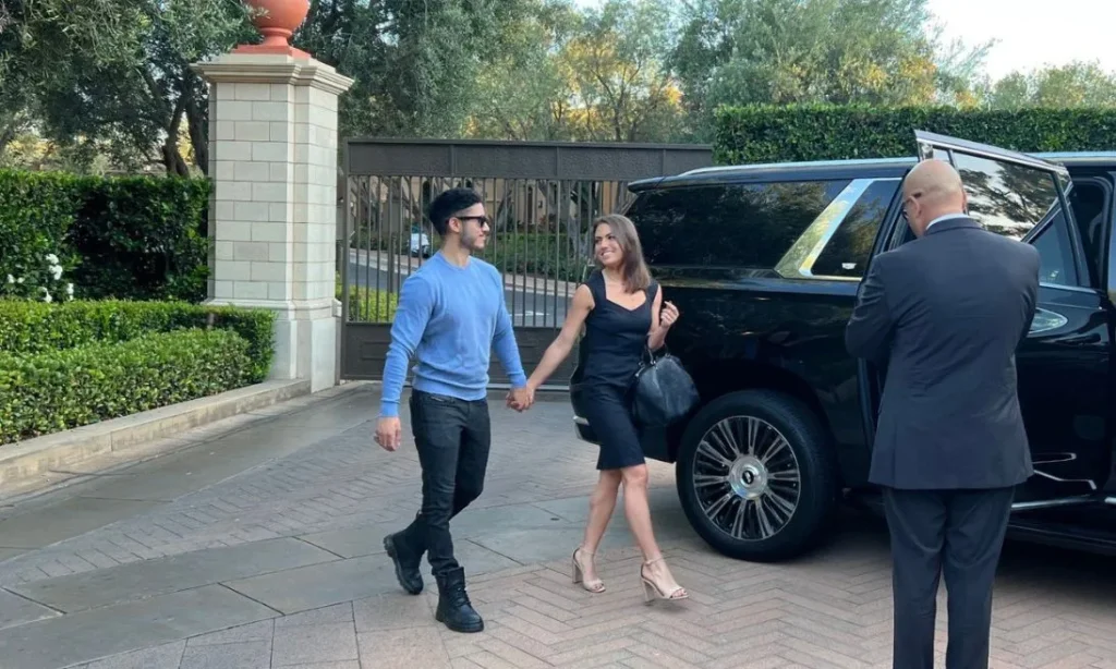 A formally dressed man and woman walk hand-in-hand toward a black SUV with an open door, as a chauffeur stands beside the vehicle.