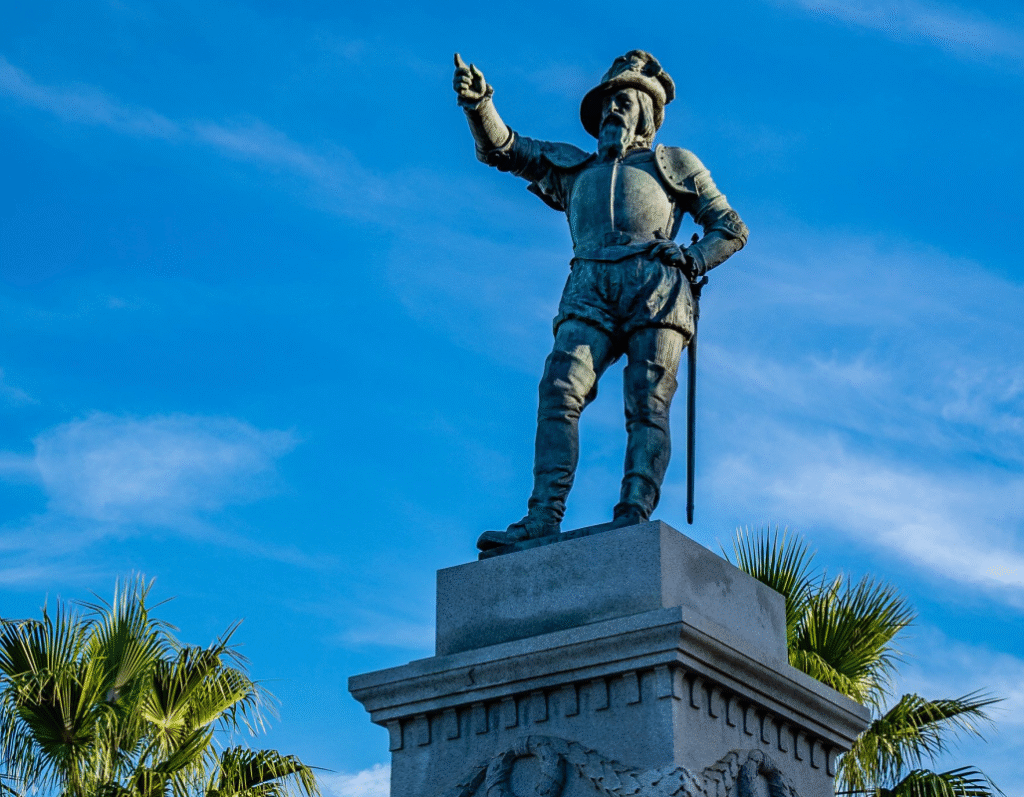 Bronze statue of Juan Ponce de León in period armor, pointing into the distance with his right hand, sword at his side, against a blue sky with scattered clouds and palm fronds in the corners.
