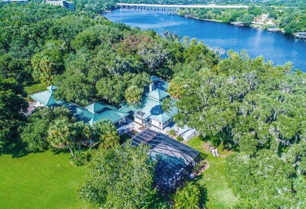 Aerial view of Riverbend Retreat, a large green-roofed building among lush trees beside a wide blue river, with a bridge in the distance; screened swimming pool and manicured lawn in the foreground.