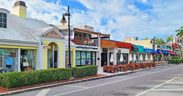 Street lined with shops and restaurants, including "Sahara" and "Hatley," featuring outdoor seating, green hedges, a streetlamp on the left, and a partly cloudy sky.