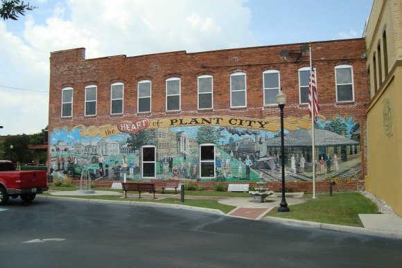 Eye-level view of red brick building in Plant City, Florida, with large mural depicting local history, including a train and townspeople; red pickup truck on the left, streetlamp and American flag on the right.