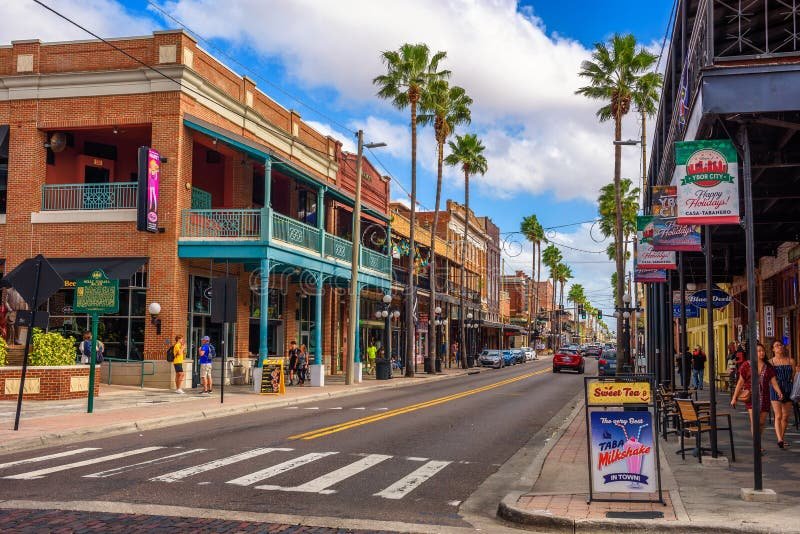 Vibrant street scene in Ybor City, Tampa, Florida, with historic brick buildings and balconies, palm-lined street, crosswalk in the foreground, pedestrians, and business signs for "Sweet Tea" and "TABA Milkshake."