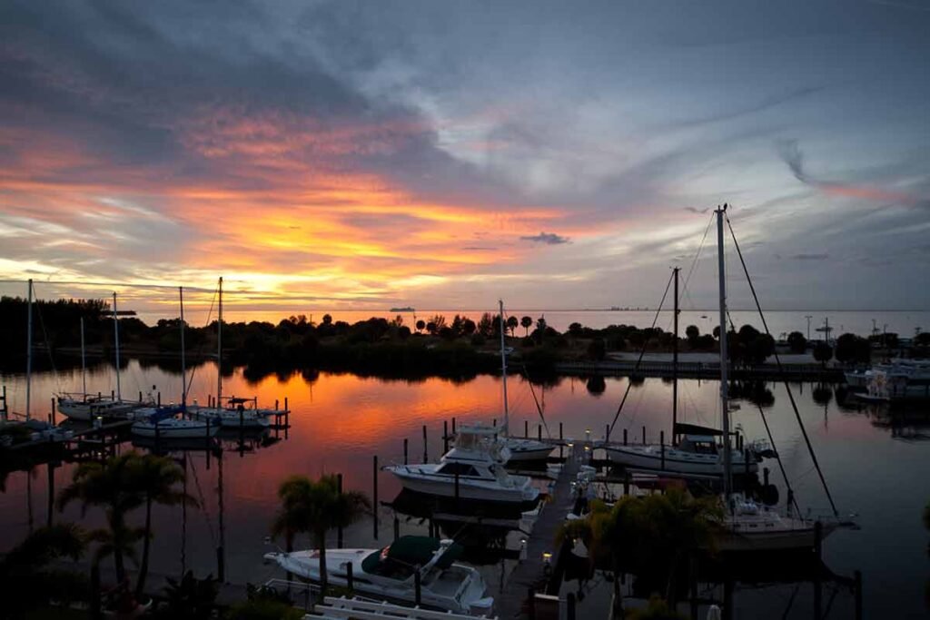 Colorful sunset over a marina with boats docked and reflected in calm water, with palm trees and distant land on the horizon.