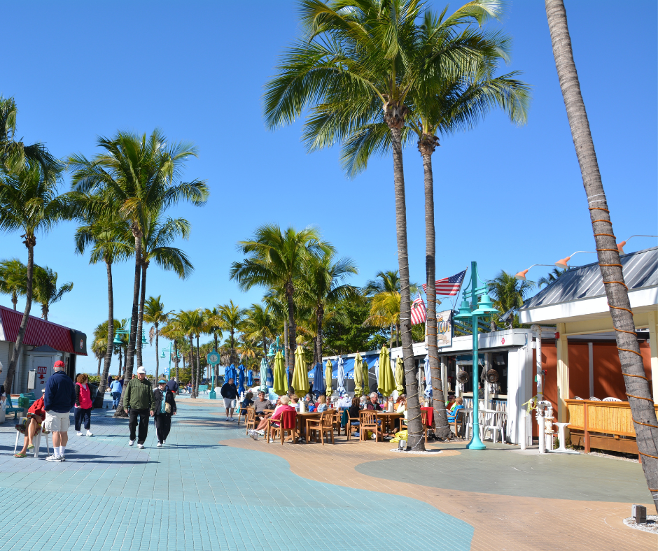 Vibrant street scene with tall palm trees and outdoor dining under umbrellas, people walking and seated at tables, all under a clear blue sky.