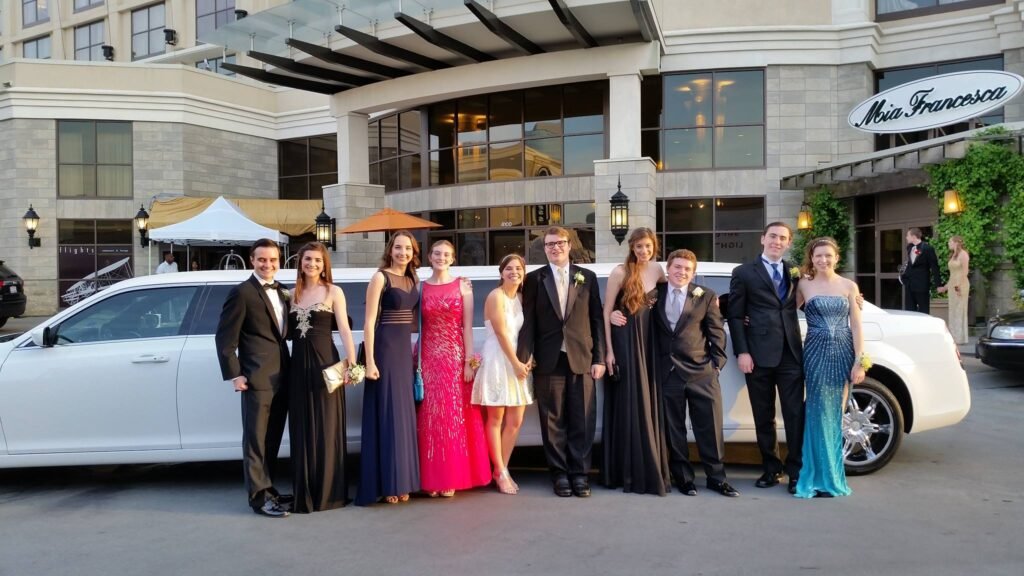 Nine young adults in formal attire pose by a white stretch limo outside a building with "Mia Francesca" sign.