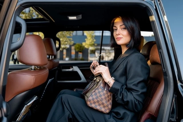 Woman with dark hair in a black suit, holding a checkered handbag, sits in a luxury car’s brown leather back seat, looking toward the viewer.