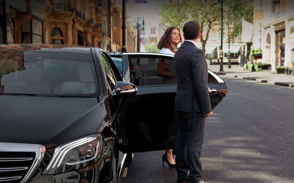 Chauffeur opening black luxury car door for female passenger on city street.